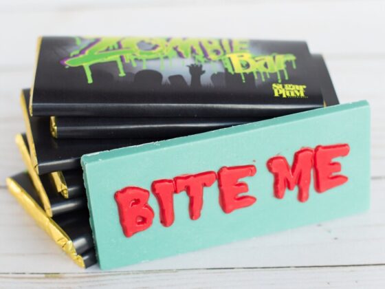 An unwrapped green zombie chocolate bar with the phrase, "BITE ME" in raised red letters, leaning against a stack of 7 wrapped candy bars.
